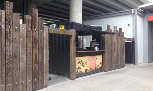 Outdoor shipping container kiosk with serving counter behind timber fence